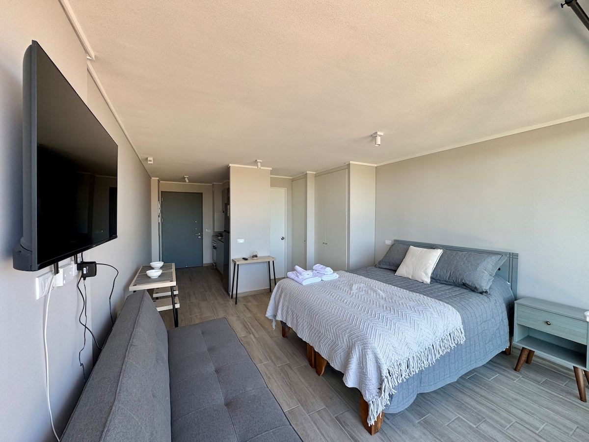 A well-lit studio features a cozy sleeping area with a spacious bed covered in soft linens and complemented by a plush futon. A wall-mounted television is positioned across from the bed, and a small dining table is visible nearby, creating a functional living space.