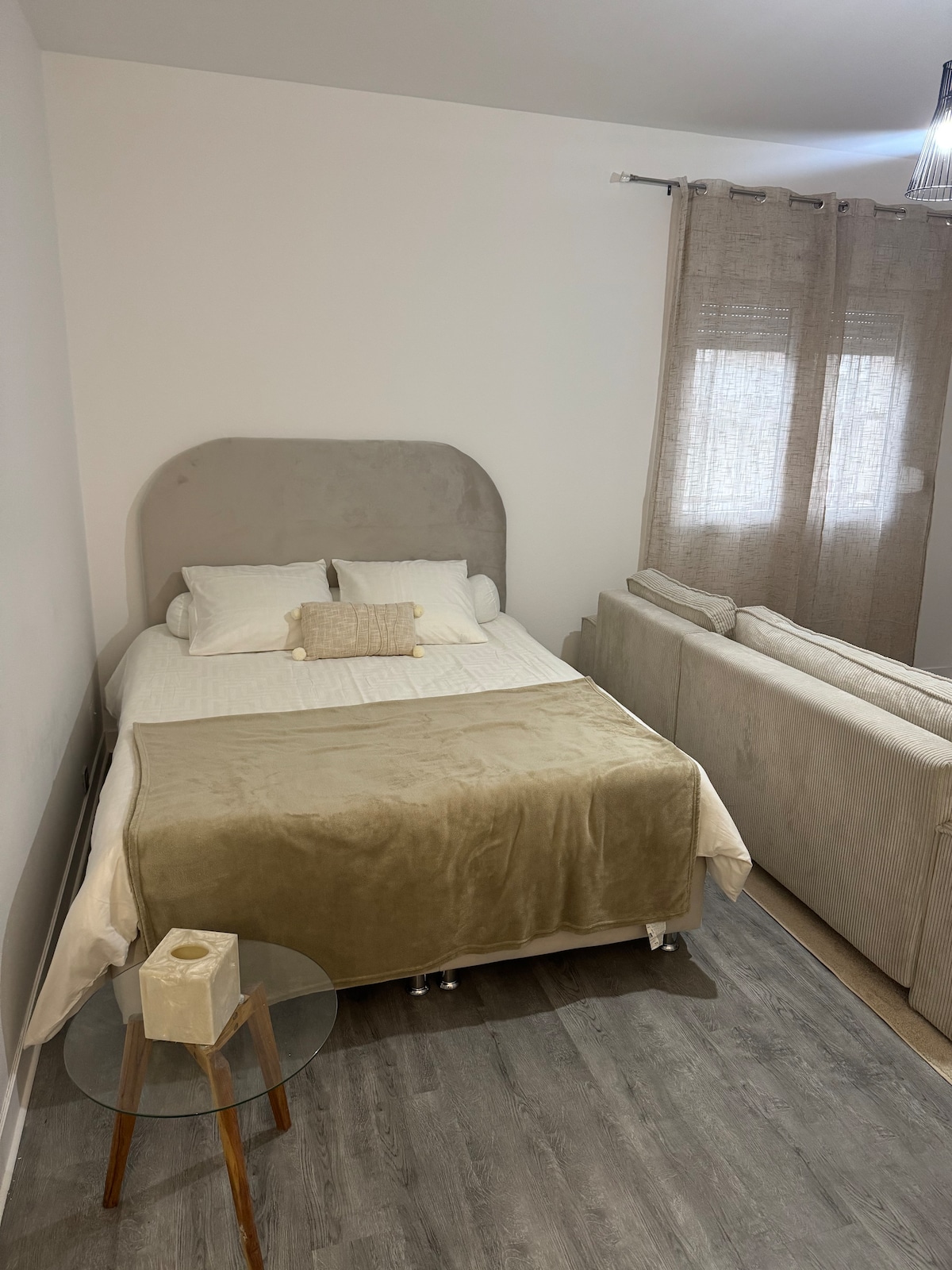 A cozy sleeping area is shown with a spacious bed, adorned with neutral-toned bedding and decorative cushions. A small side table is positioned beside the bed. A soft-textured sofa is visible in the background, complemented by sheer curtains allowing natural light to filter in.