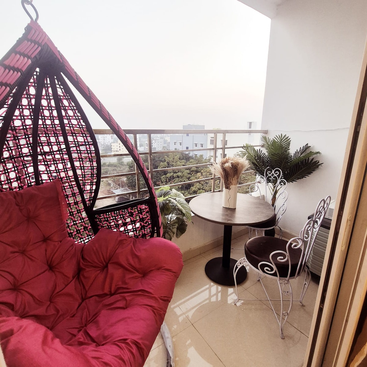 A cozy balcony features a hanging chair with red upholstery and a small round table beside two white wrought-iron chairs. Potted plants are visible, adding greenery to the space, while the expansive view showcases the surrounding cityscape.