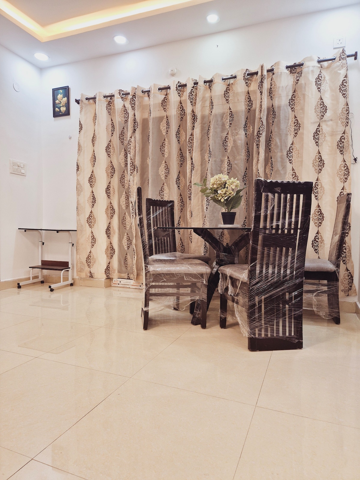 A dining area is presented with a round table surrounded by four dark wooden chairs. A vase with flowers is centered on the table. Sheer curtains are seen covering a window, while a small side table is placed nearby. The floor is tiled and reflects light.