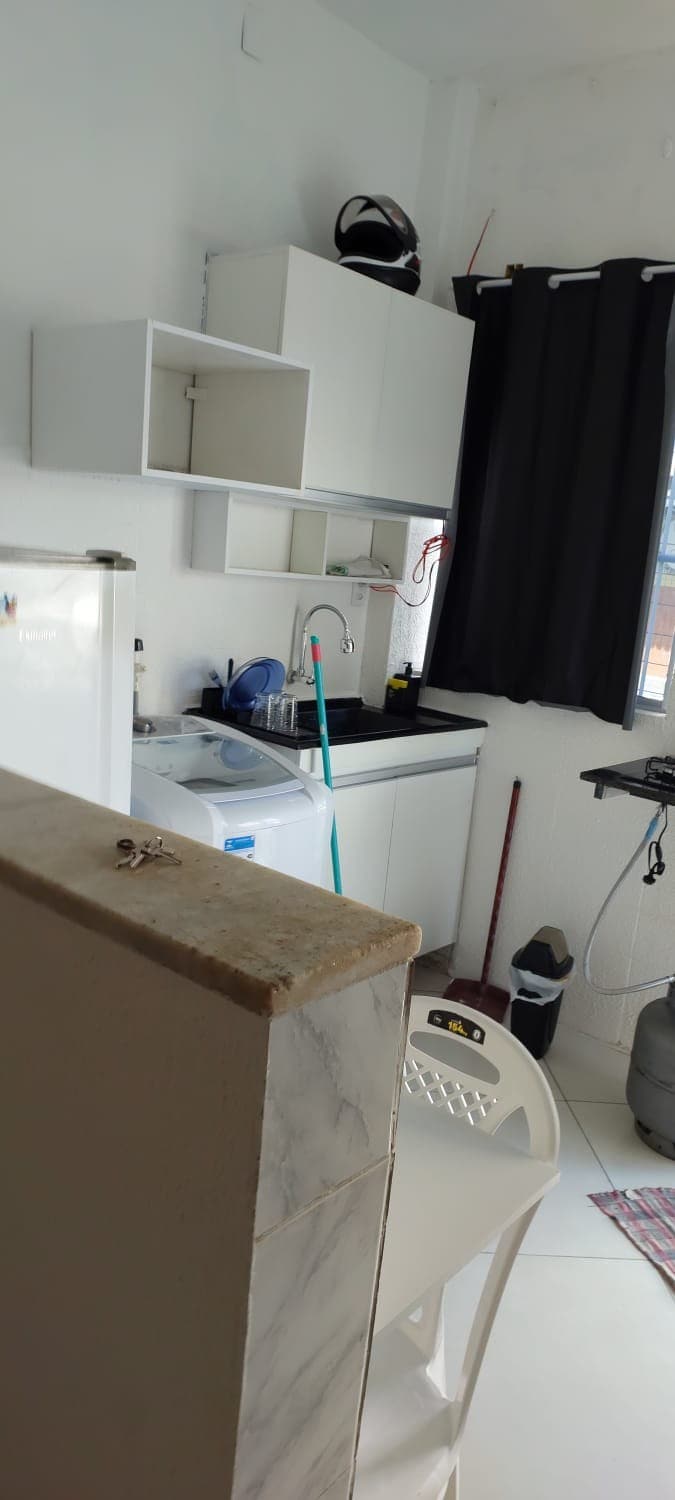 A functional kitchenette features a compact refrigerator and a washing machine, along with a sink and overhead cabinets. A black countertop provides workspace, while a window is dressed with dark curtains. A small white chair and cleaning supplies are also visible.