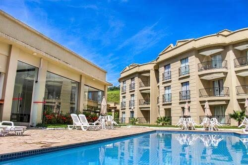 A well-maintained pool area is visible, surrounded by lounge chairs and umbrellas. Two adjoining buildings feature multiple balconies, each with a distinct architectural design. Lush greenery complements the serene atmosphere under a clear blue sky.