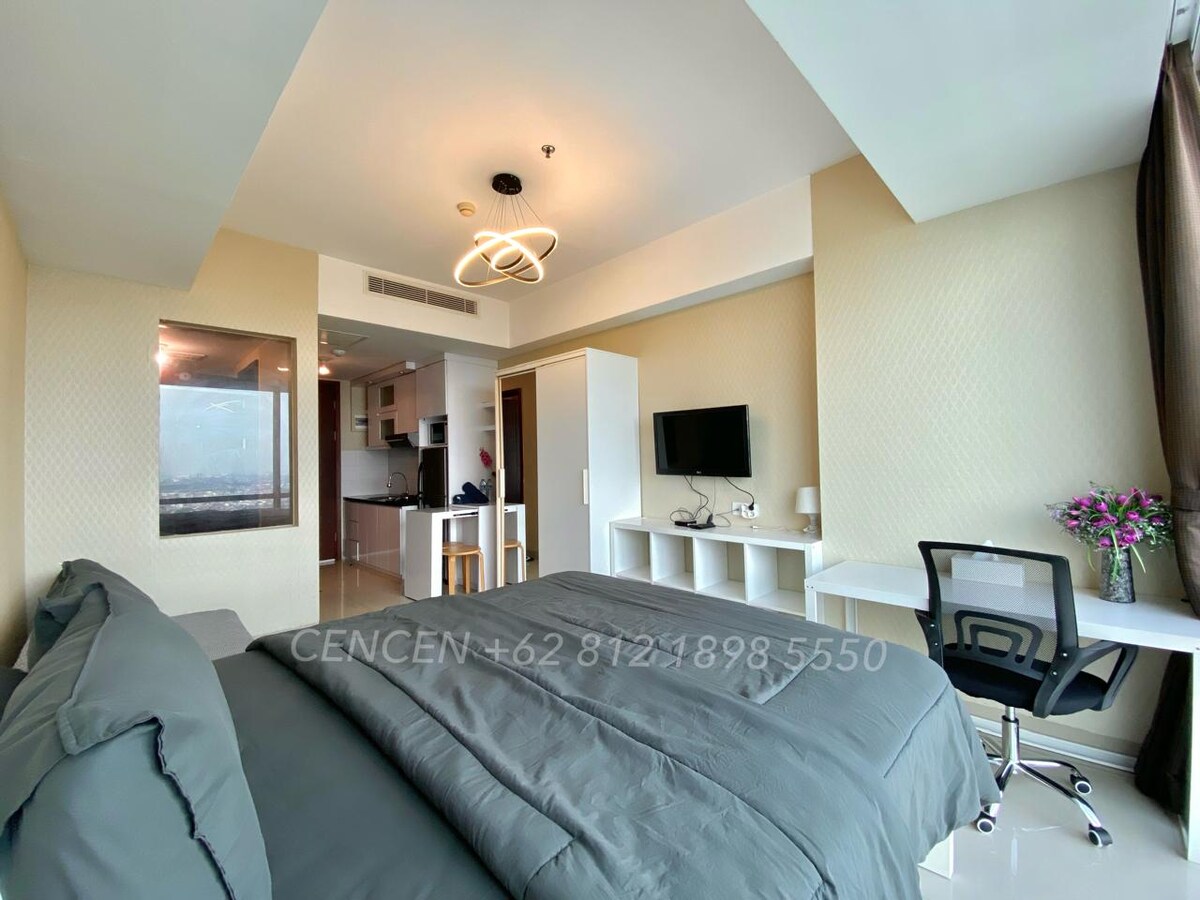 A spacious bedroom is equipped with a large bed featuring neutral-toned bedding. A flat-screen TV is mounted on the wall across from the bed. A desk with a simple chair is positioned by a window, allowing for natural light to fill the space.