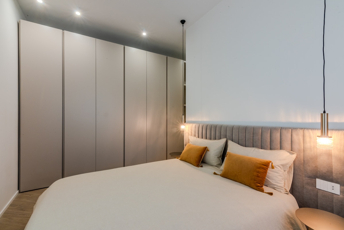 The spacious bedroom features a comfortable bed dressed in neutral tones, complemented by two decorative pillows. Ample built-in wardrobes stretch along one wall, while soft lighting is provided by pendant lights on either side of the bed, adding a cozy touch.