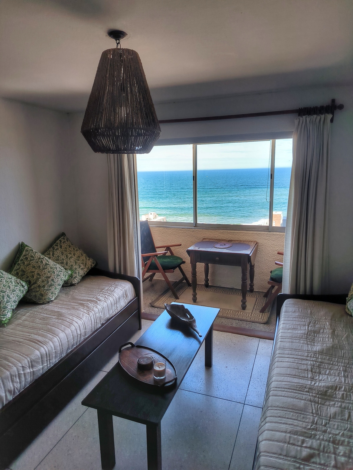 The living area is furnished with two twin beds and a dark wood coffee table, providing a cozy setting. A large window offers an expansive view of the ocean, while natural light fills the room. Decorative pillows add a touch of comfort to the seating.