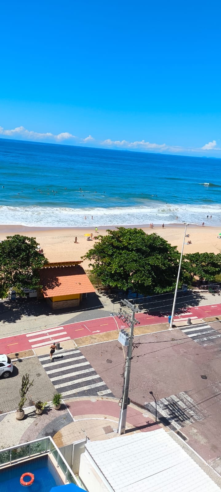A view of the beach reveals soft sandy shores meeting the ocean. Lush green trees line the beachfront, while people enjoy the sun and water. In the foreground, a crosswalk and road can be seen, indicating the proximity to urban amenities.