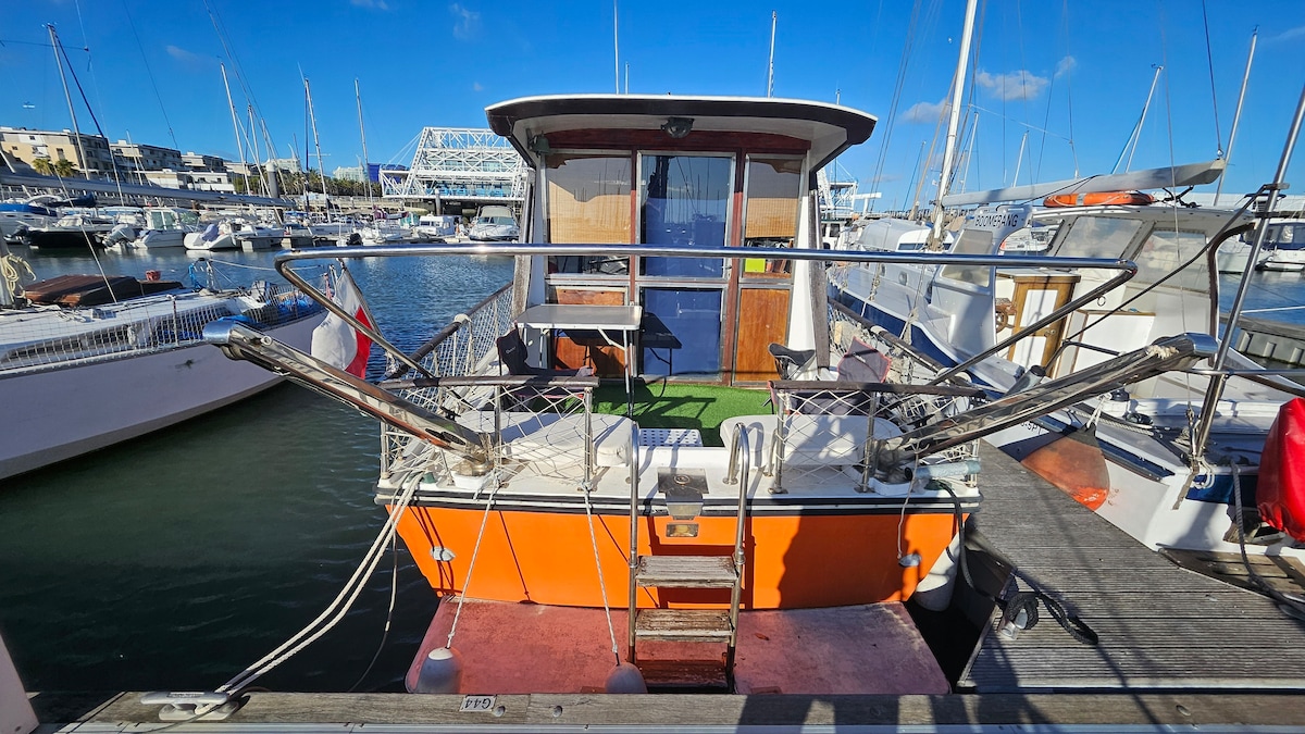 The rear view of the boat displays an orange hull with a wooden deck. Two railings are visible, leading towards the entrance. The marina backdrop includes various boats and yachts docked nearby, under a clear blue sky.