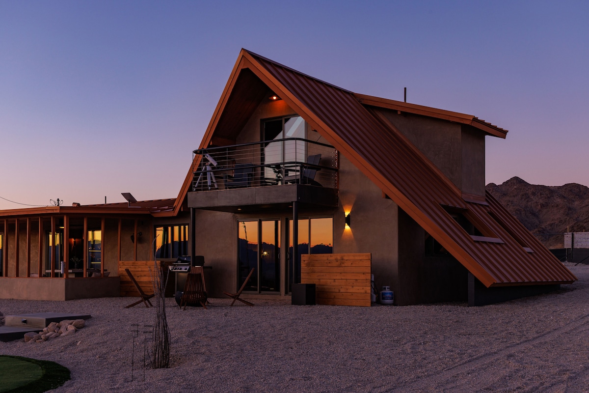 An A-frame retreat features a striking triangular roof and is surrounded by desert landscape. The exterior is crafted with earthy tones and large windows, allowing natural light to flood the interior. Relaxation areas and outdoor seating are visible, enhancing the serene atmosphere.