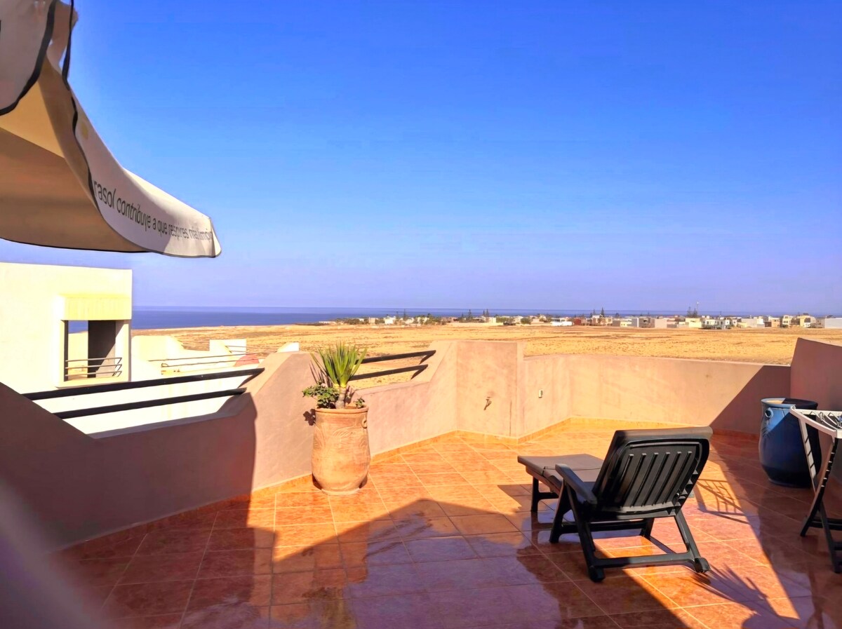 A spacious terrace is shown with a partial sea view, complemented by a view of the mountains in the background. A single lounge chair is positioned on the tile flooring, alongside a potted plant. A shaded area is also visible from the retractable awning.