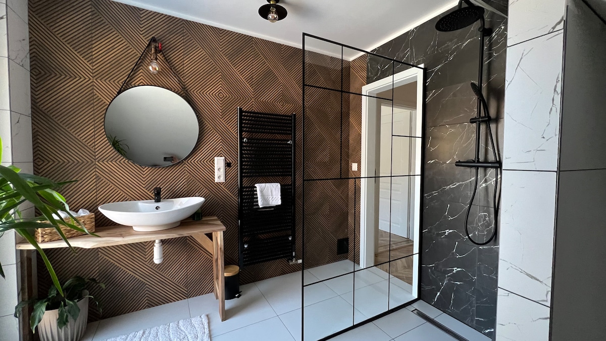 A modern bathroom features a spacious walk-in shower enclosed by clear glass. A circular mirror hangs above a minimalist sink, with wooden accents on a console table. A towel rack and potted plant add functional and decorative touches to the space.