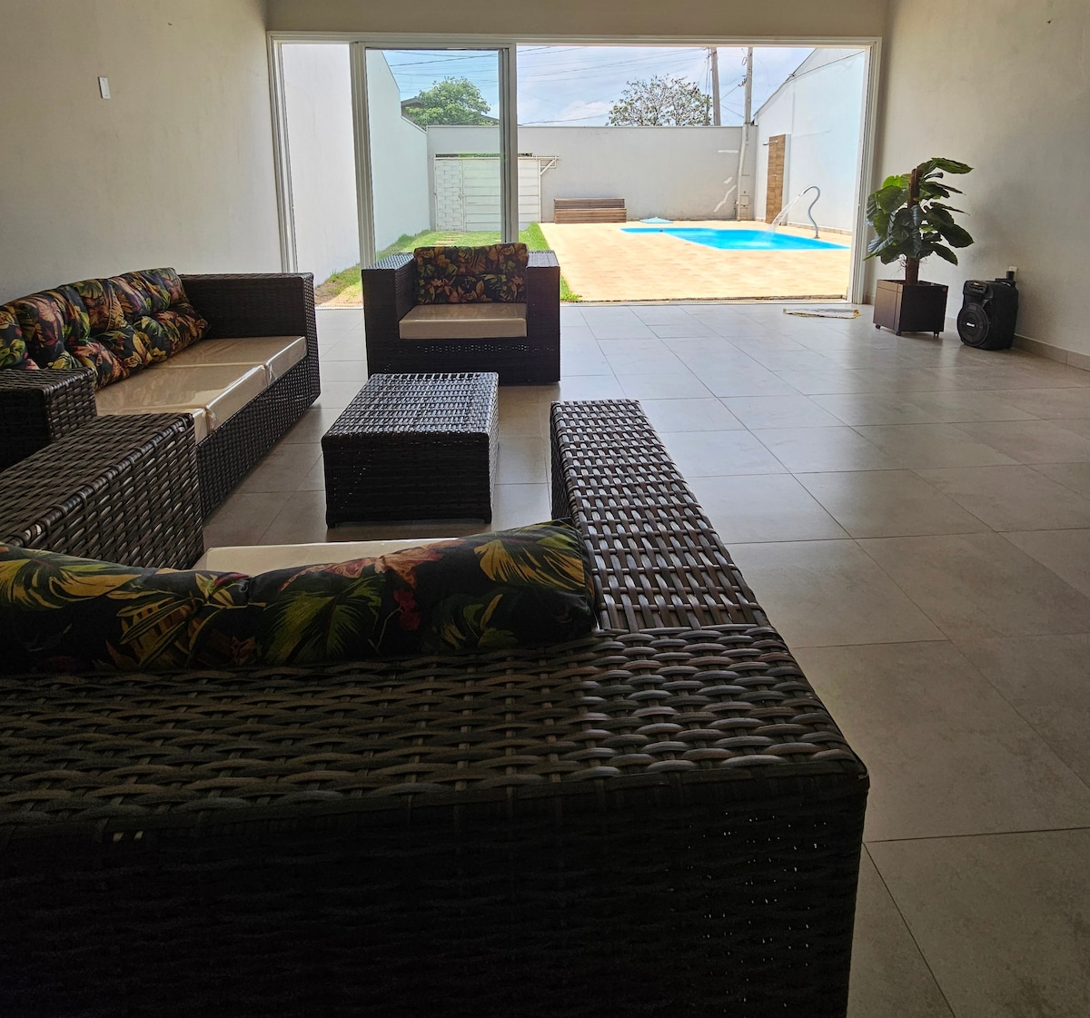 A comfortable seating area features woven furniture with colorful cushions, facing an expansive glass door. Natural light illuminates the space, creating a connection to the outdoor area, which includes a refreshing pool and a patio area in the background.