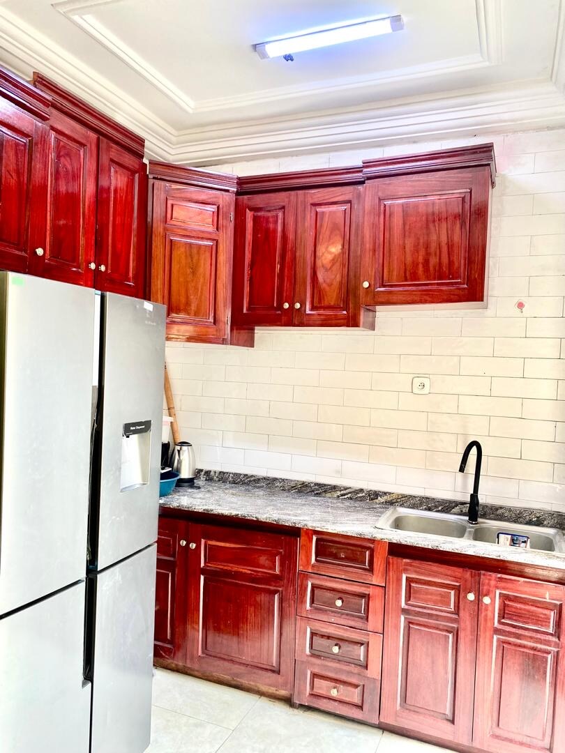 A modern kitchen features rich wooden cabinetry with a dark granite countertop. Stainless steel appliances, including a refrigerator and sink with a black faucet, are present. White tiled walls enhance the bright atmosphere, and under-cabinet lighting offers additional illumination.