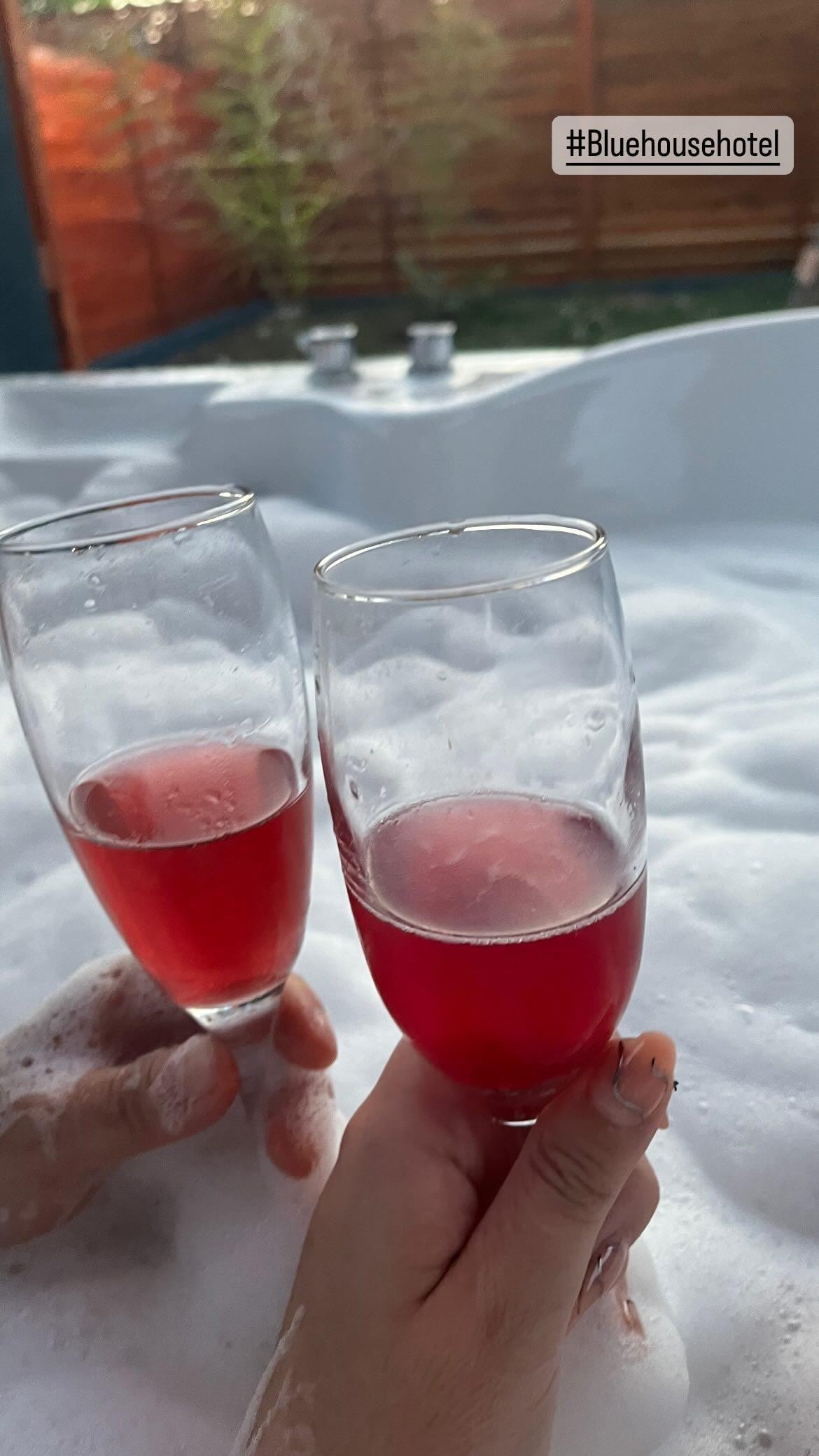 Two glasses of sparkling red beverages are held above a bubbly jacuzzi. Foam fills the space, creating a relaxing atmosphere. Bamboo plants are discreetly visible in the background, enhancing the serene setting.