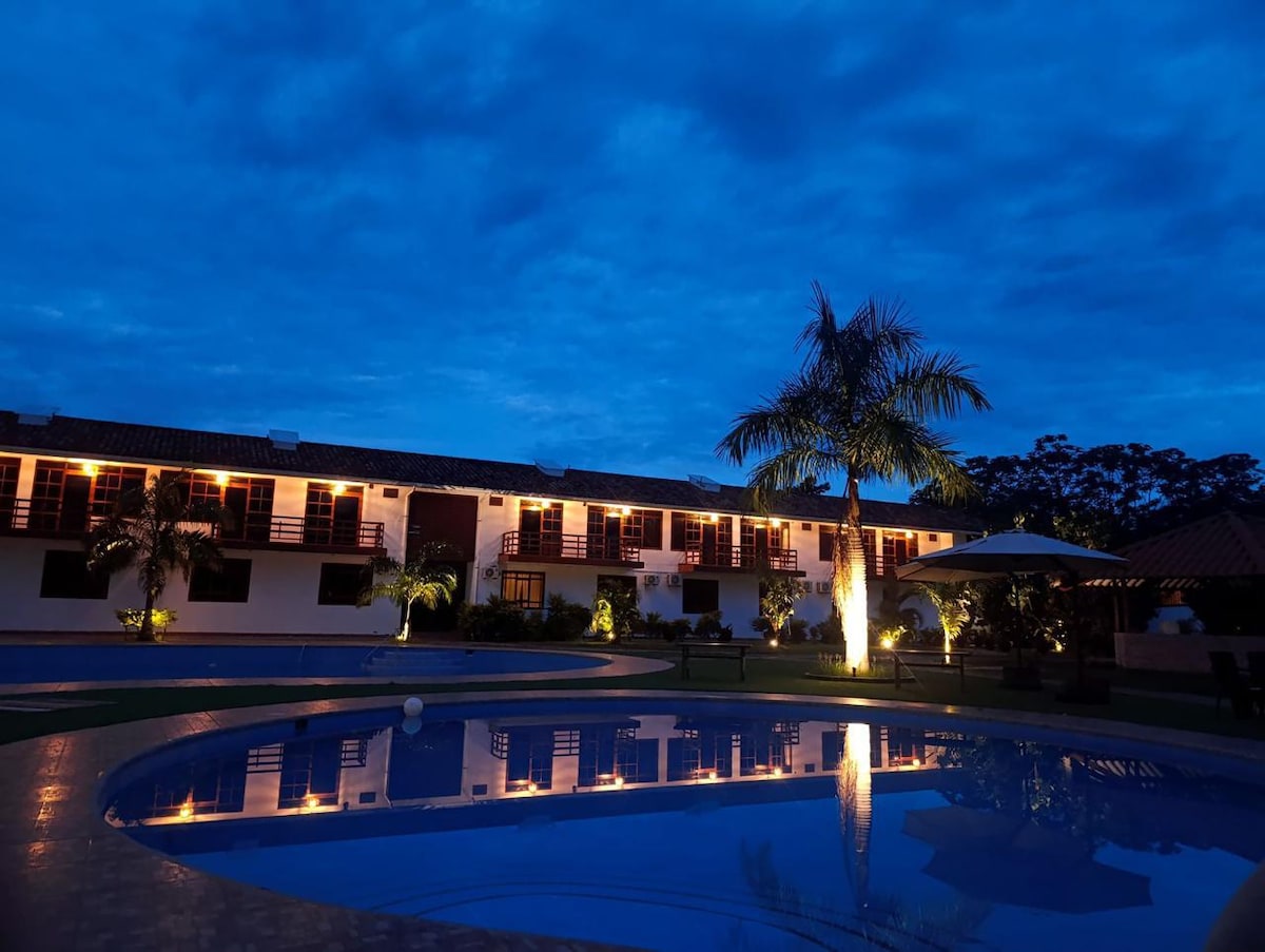 The tranquil nighttime setting features a well-lit pool area surrounded by palm trees. The bungalow's facade is visible, with warm lights illuminating the building's exterior. The calm water reflects the cloudy sky, creating a serene atmosphere.