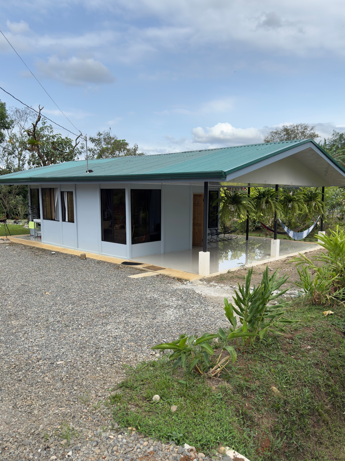 A single-story dwelling is presented with a sloped green roof and large windows that allow natural light to enter. Surrounding greenery adds a sense of tranquility, while a hammock is visible on the porch, inviting relaxation in the outdoor space.