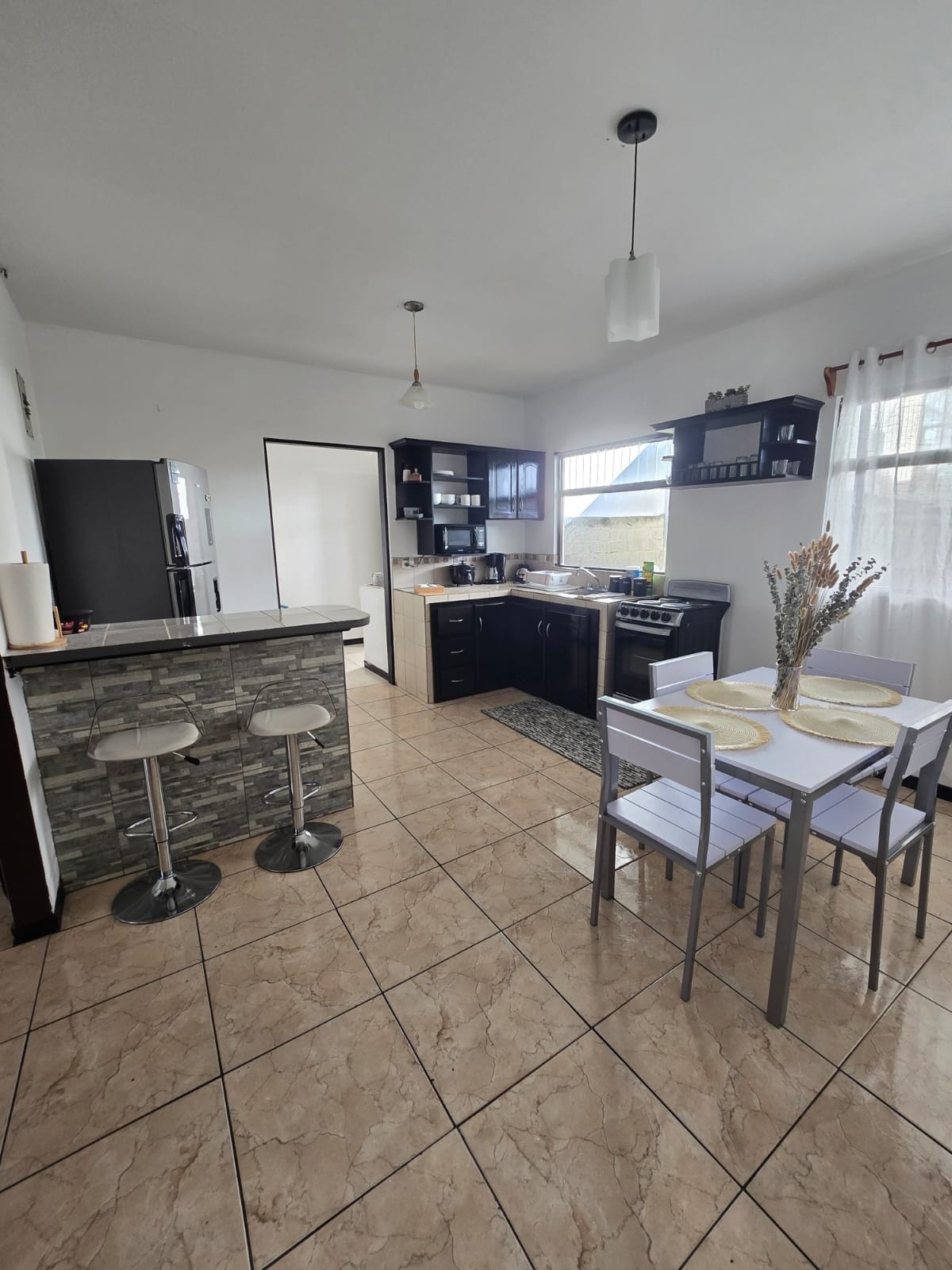 A modern kitchen is highlighted by dark cabinets and a light countertop, featuring an organized layout. A dining area is positioned near a window, with a small table and four chairs. Bar stools are situated at the counter, enhancing the space's functionality.