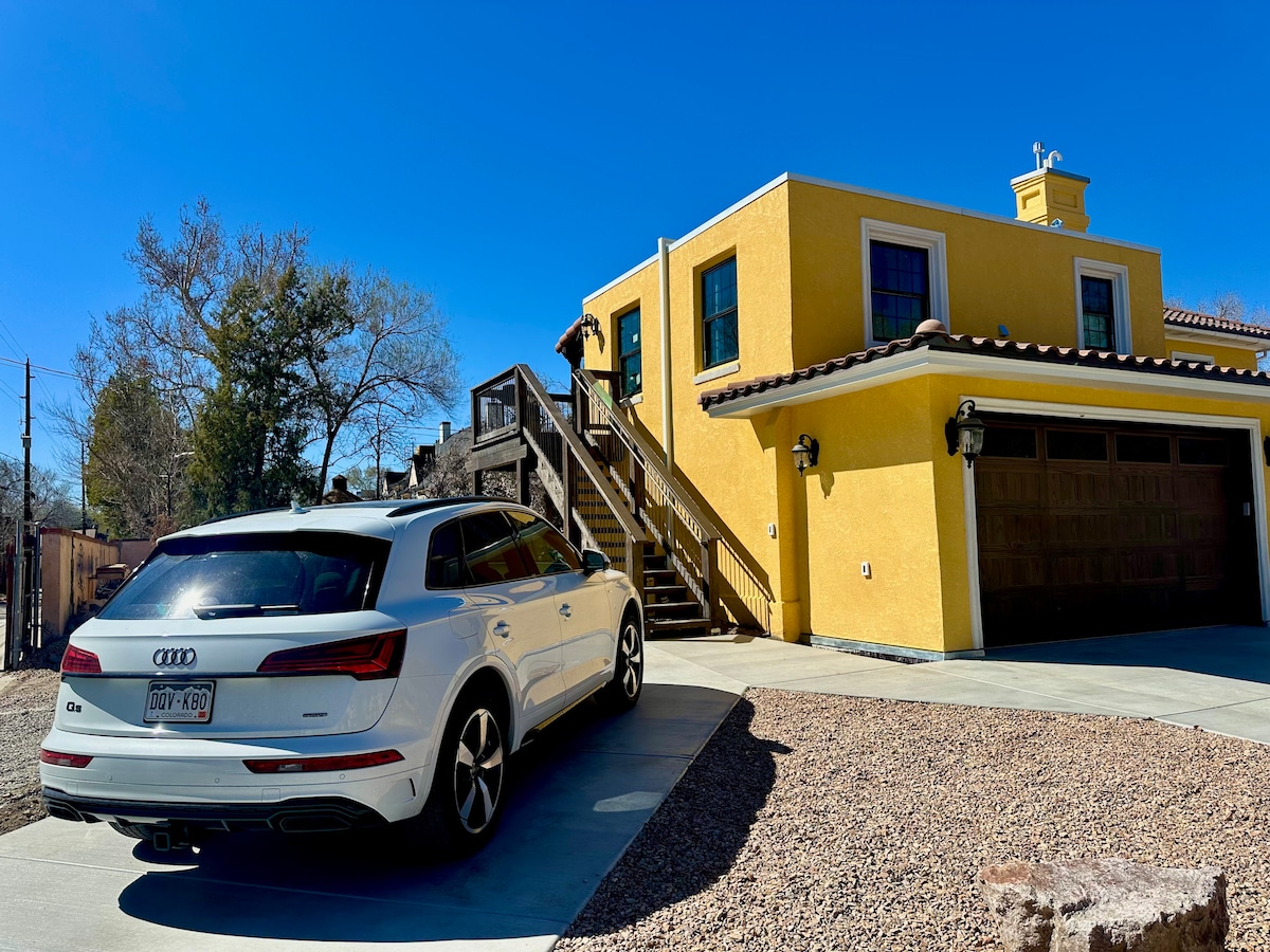A bright yellow building with windows and a staircase leading to the entrance is shown. A white vehicle is parked in the driveway, surrounded by gravel and a stone border. Clear blue skies and trees are visible in the background.