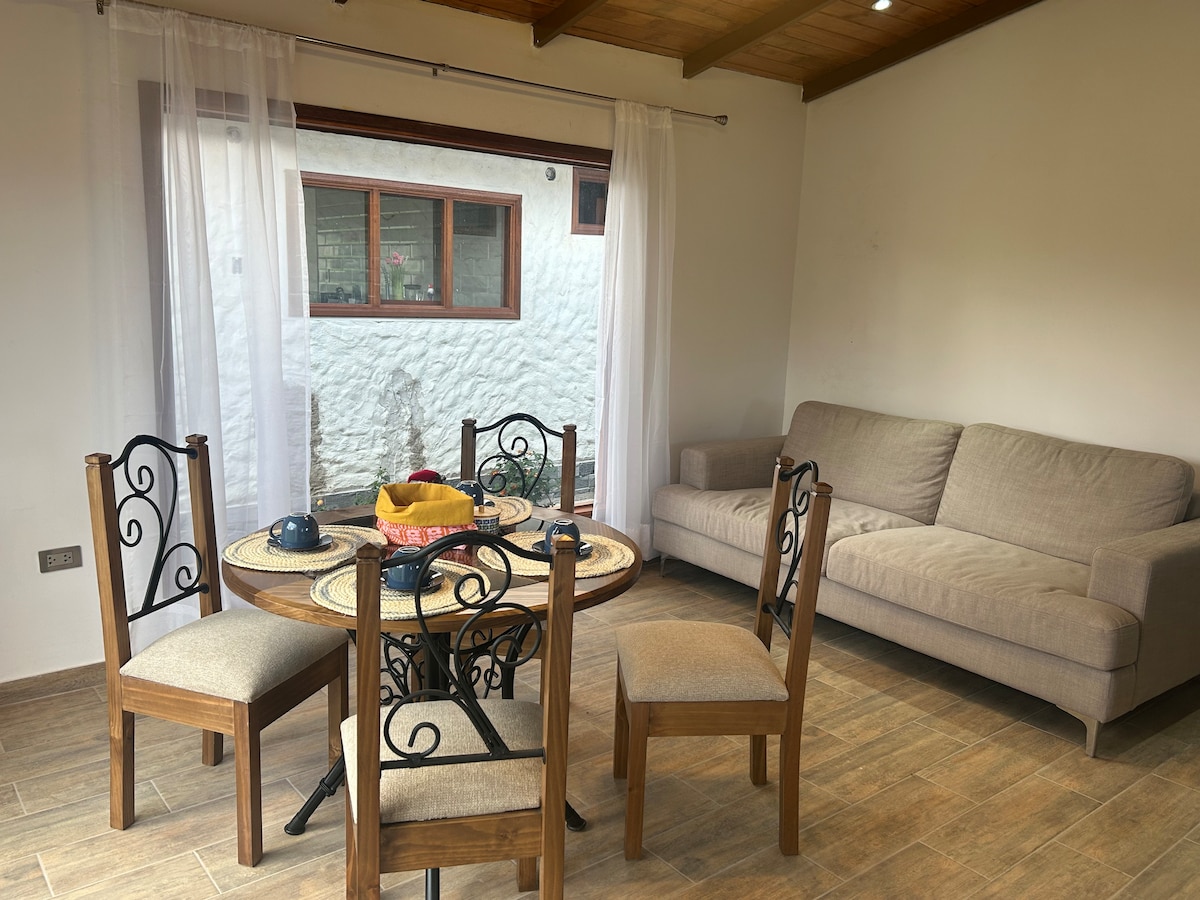 A dining area features a round wooden table surrounded by four chairs. A sofa is placed against the wall, and soft curtains frame a window that allows natural light to enter, revealing a glimpse of the outdoor space.