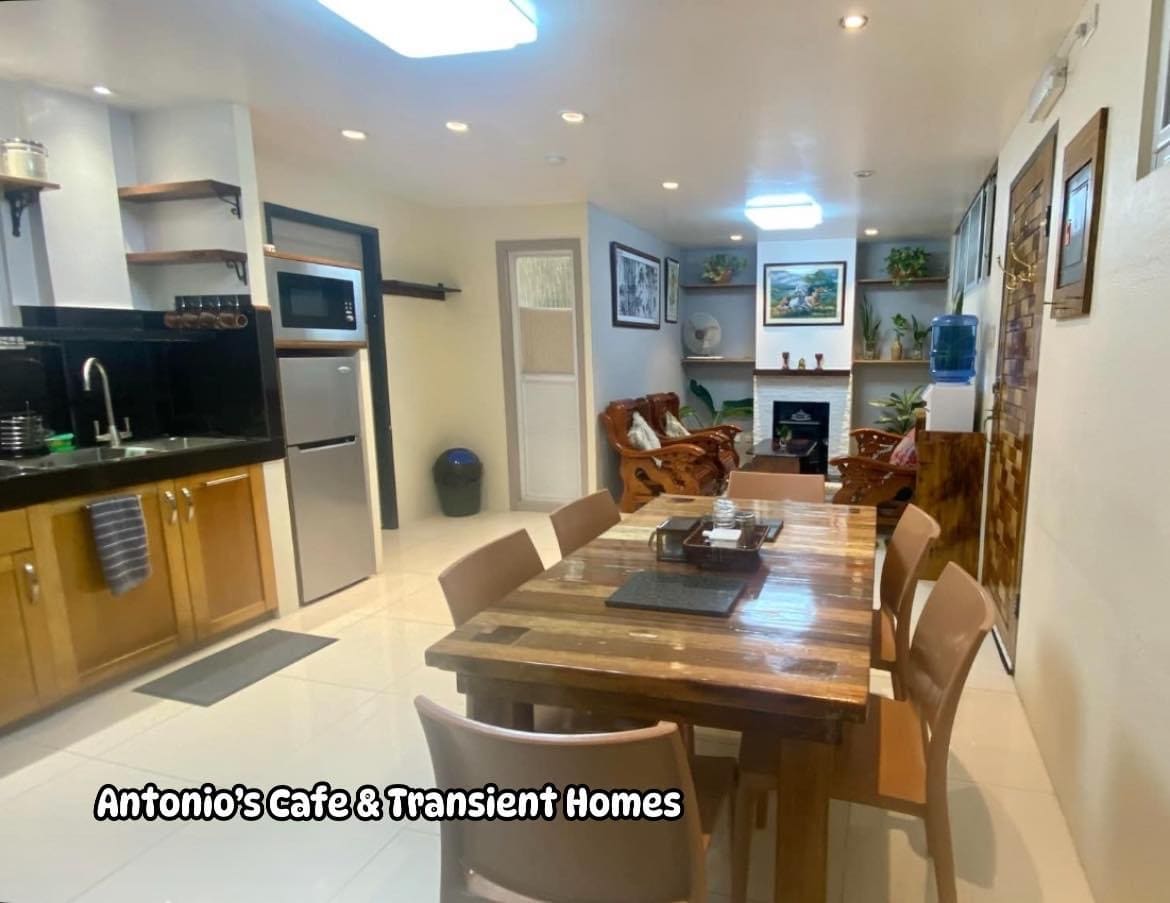 A modern kitchen area is visible, featuring wooden cabinets and stainless steel appliances, including a refrigerator. A six-seat dining table, made of natural wood, is positioned centrally. A cozy seating area with armchairs and framed artwork is seen in the background, enhancing the inviting space.