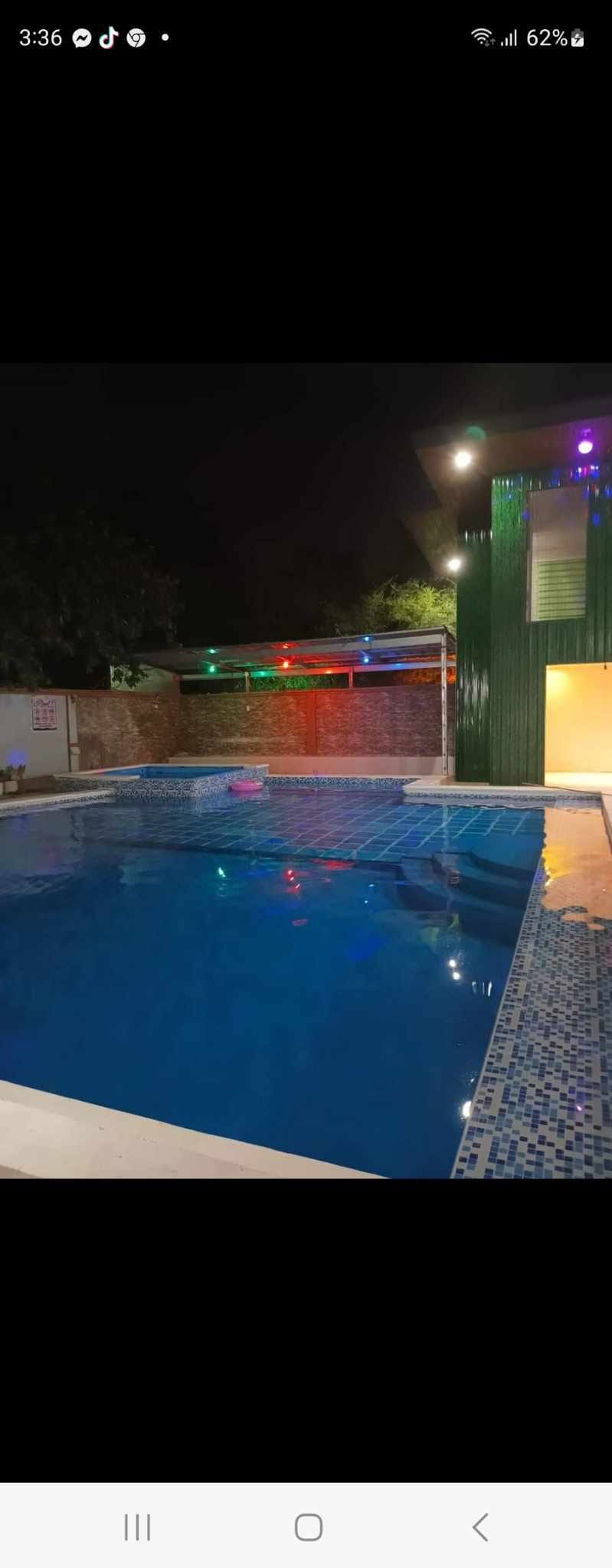 A well-lit swimming pool is framed by colorful lights, enhancing the evening ambiance. The pool features a clear blue surface, reflecting the surrounding area. A brick wall provides a backdrop, while green structures and trees are visible in the distance.