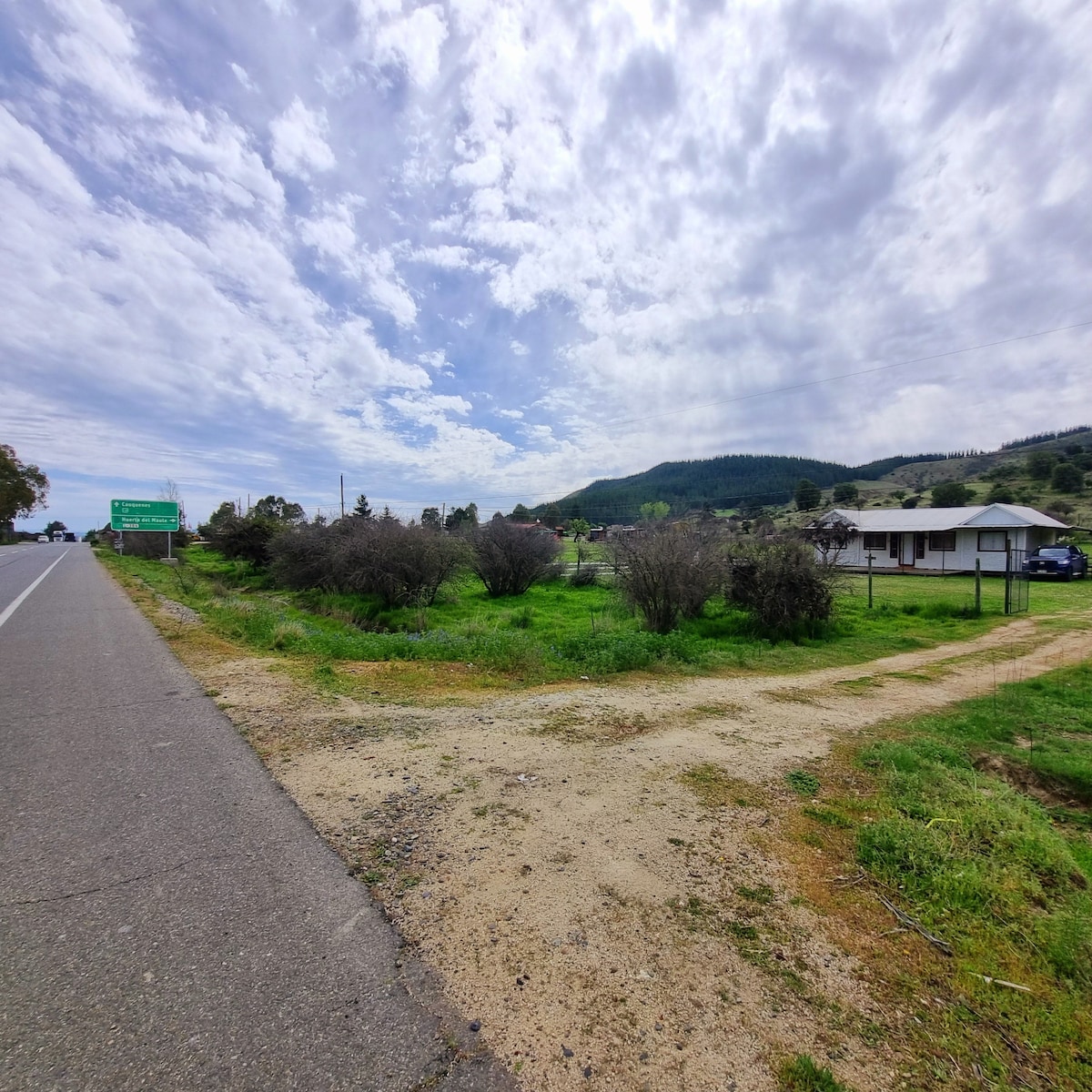 A view of a serene landscape is presented, featuring lush green grass and shrubs along a dirt pathway. A modest building is visible to the right, surrounded by open space, with distant hills and a cloudy sky creating a tranquil backdrop.