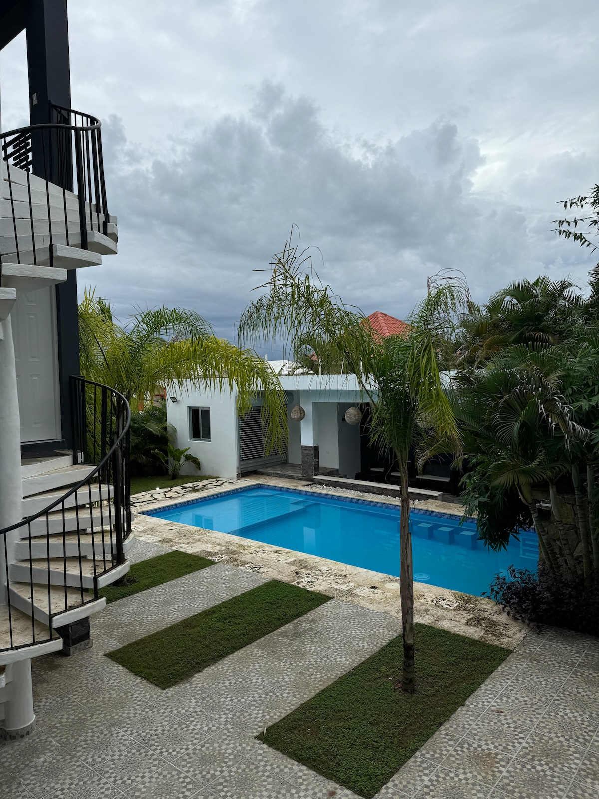 A swimming pool is situated amidst tropical landscaping, featuring palm trees and manicured lawns. The pool area is bordered by a mosaic-tiled patio, offering ample outdoor space. A modern two-story villa is visible, with a spiral staircase leading to the upper level.