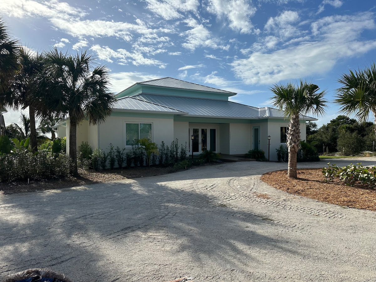 A modern single-story house is situated amidst palm trees and native vegetation. The structure features a light exterior with a metal roof, and a spacious driveway leads to the entrance, where a welcoming atmosphere is created by the surrounding natural landscaping.