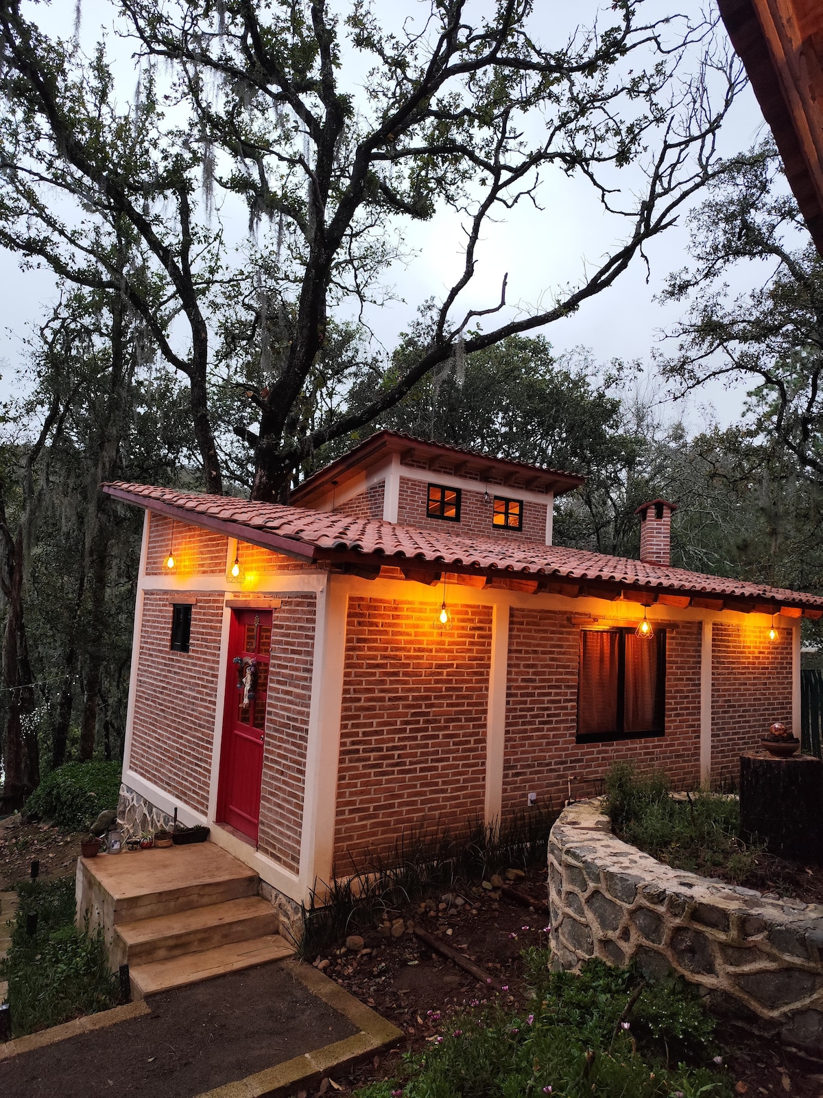 A welcoming cabin is surrounded by lush trees, featuring a distinct red door and warm outdoor lighting. The brick exterior is complemented by a terracotta roof. Steps lead up to the entrance, and a stone wall outlines the property, enhancing the natural setting.