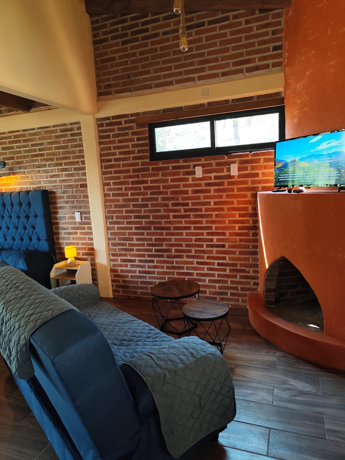A seating area features a plush blue sofa facing a modern television set atop a circular black table. Exposed brick walls accentuate the warm atmosphere, while a cozy fireplace is integrated into the design, providing a focal point within the space.