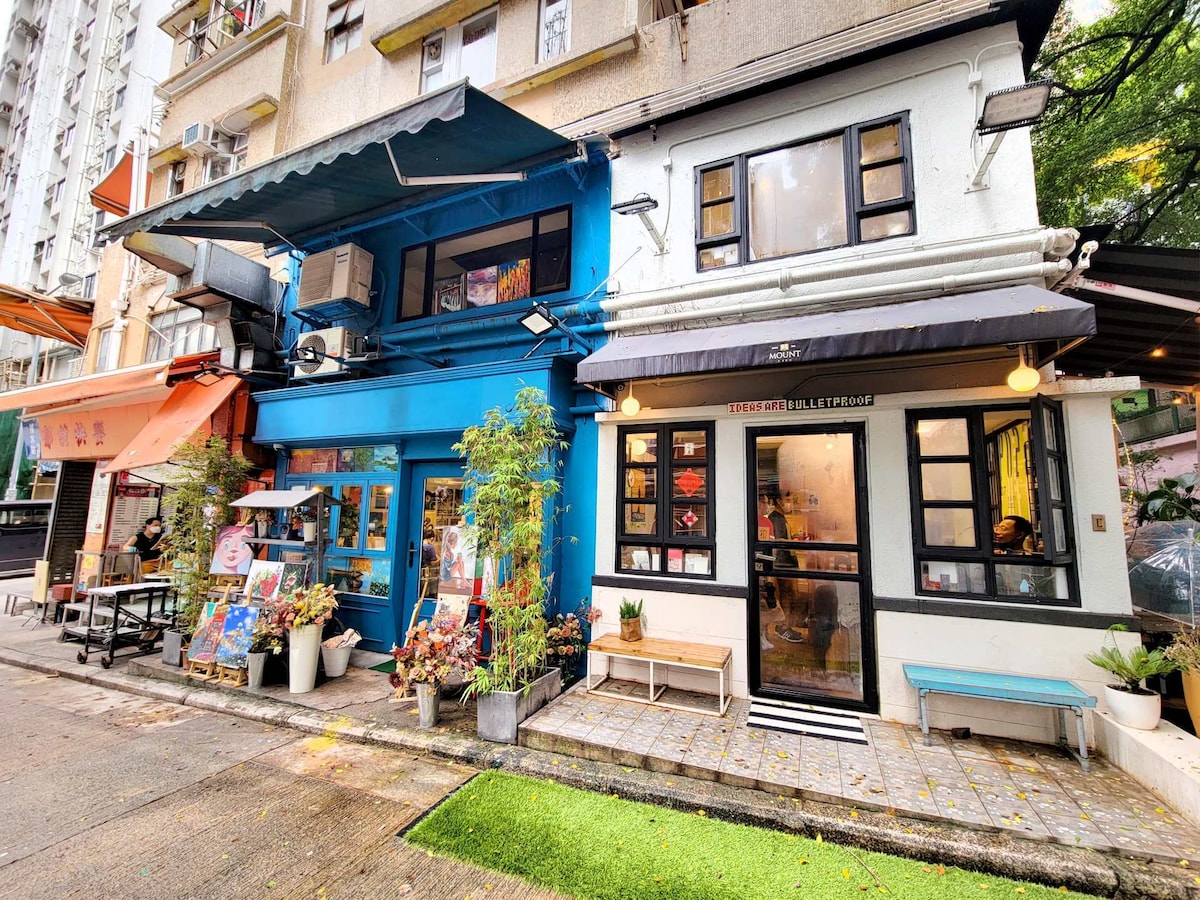 A colorful row of buildings showcases an inviting street scene. The blue and white structures highlight large windows and awnings. Planters with greenery line the walkway, while outdoor seating is visible in front of the shops, creating a lively atmosphere typical of the neighborhood.