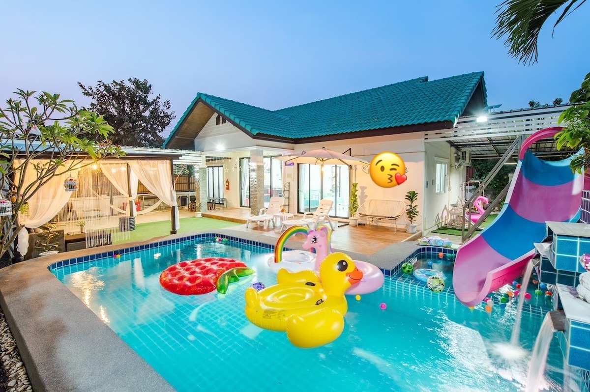 A private outdoor pool features colorful inflatable floats, including a duck and a large slice of watermelon. Surrounding the pool are lounge chairs and vibrant greenery, with a covered area providing additional seating options. The house is set against a twilight sky, enhancing the inviting atmosphere.
