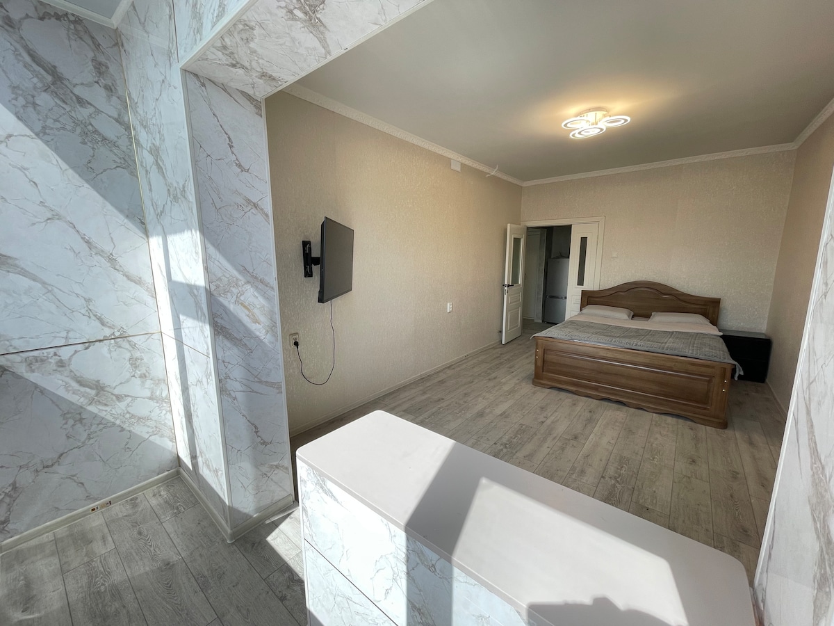 A spacious bedroom features light-colored walls and wood flooring. A large wooden bed is positioned centrally, accompanied by a flat-screen television mounted on the wall. Natural light enters through a nearby window, illuminating the room's minimalist design.