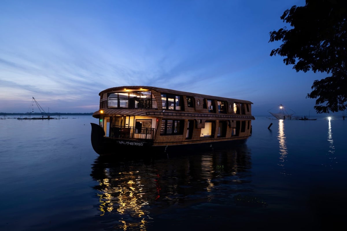 A traditional Kettuvallam boat is anchored gently on calm waters, illuminated by soft evening light. Its bamboo-thatched structure showcases multiple windows and a spacious deck, creating a serene atmosphere against a vibrant twilight sky.