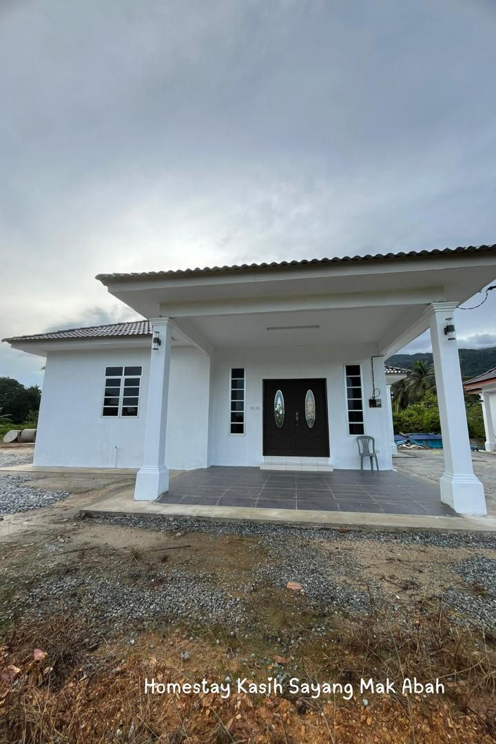 The exterior view of the homestay features a simple white façade with a covered porch. Two elegant double doors serve as the entrance, framed by two windows on each side. The surrounding area contains gravel and a landscaped environment, with mountains visible in the background.