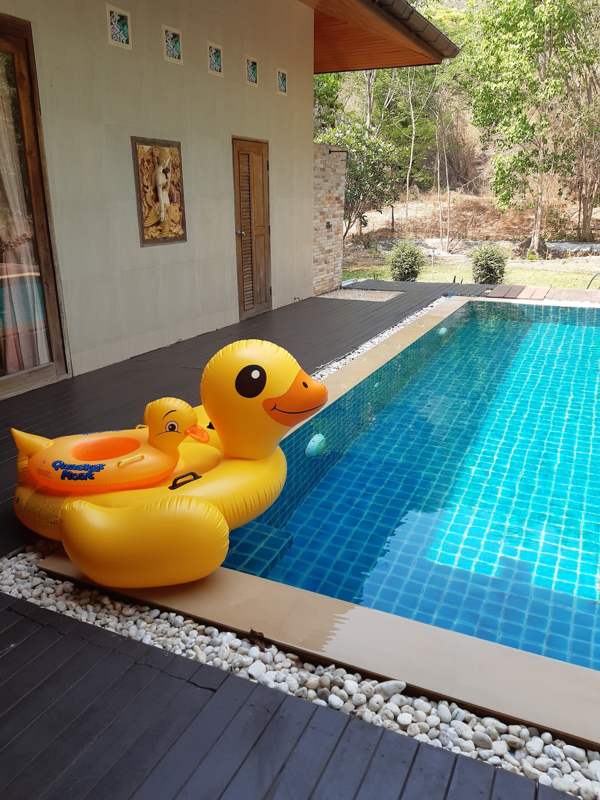 An oversized yellow rubber duck floats beside a private swimming pool, which is surrounded by smooth stones. Sunlight reflects off the clear blue tiles, while lush greenery and trees are visible in the background, enhancing the tranquil setting.