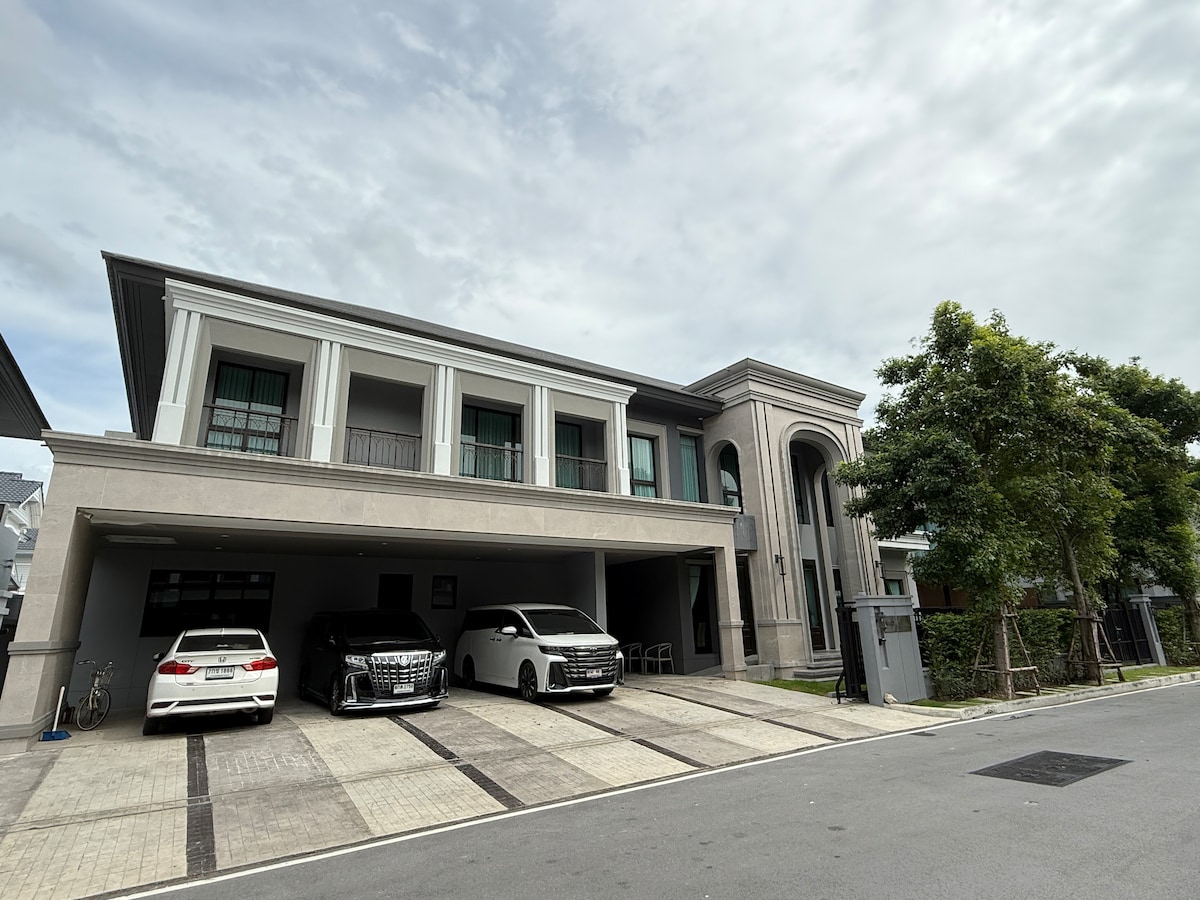 The exterior of a modern home is displayed, featuring a spacious driveway accommodating multiple vehicles. The building's architecture includes large windows and a covered porch area. Lush greenery is visible on the side, contributing to the overall spacious and inviting layout.
