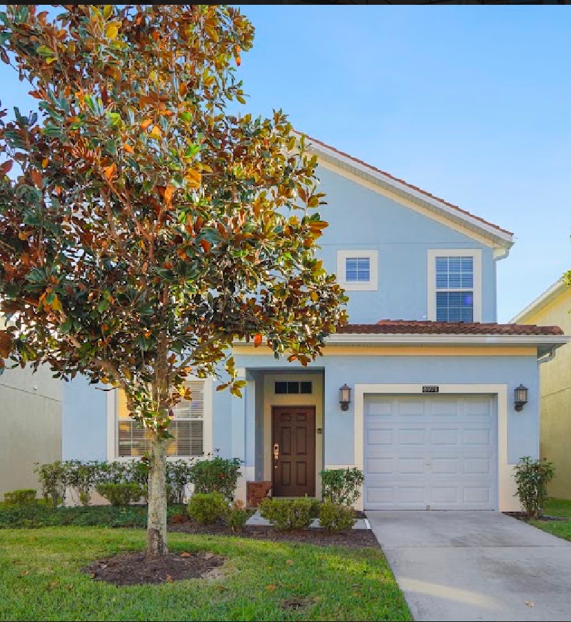 A light blue two-story home is shown, featuring a welcoming front door with a symmetrical window above it. A manicured lawn with shrubs borders the walkway, and a small tree adds greenery to the entrance. A single-car garage is visible on the left.