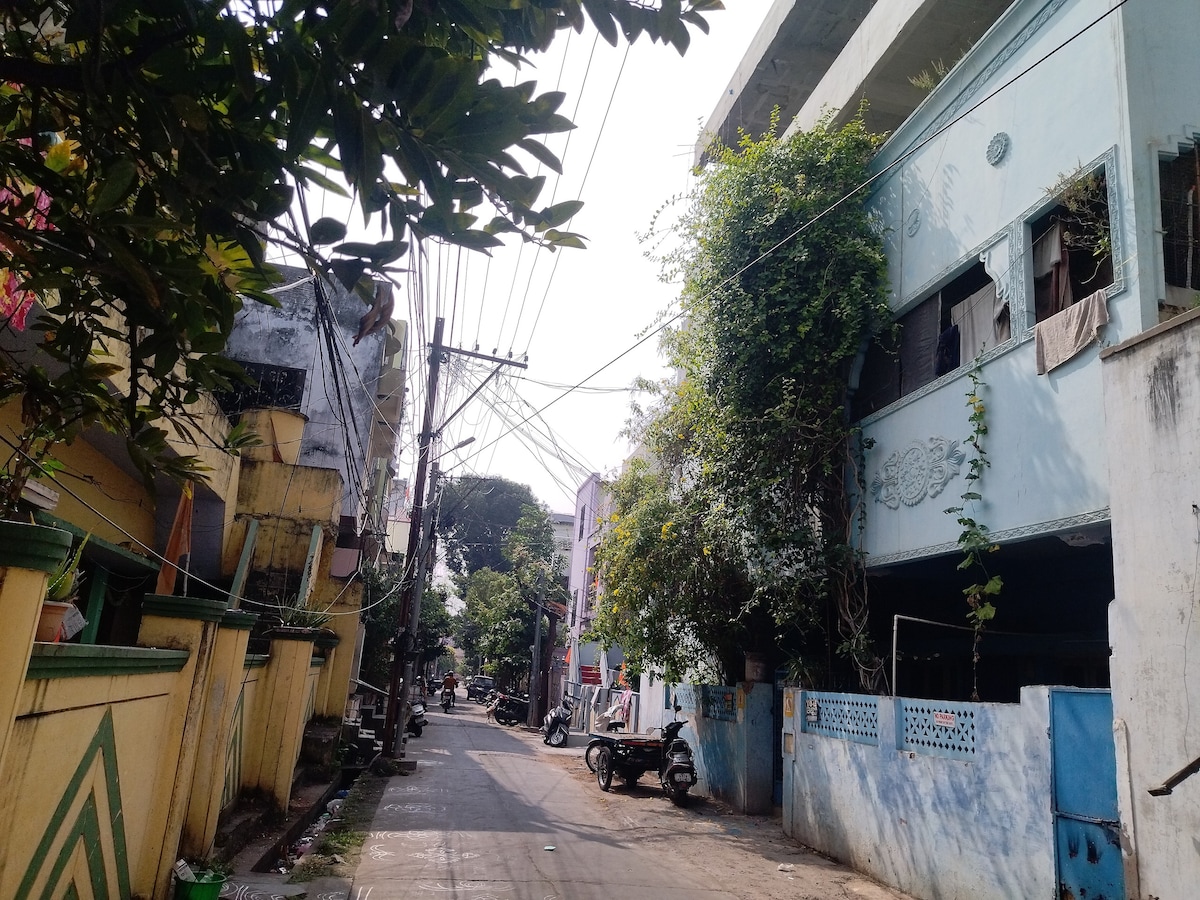 A quiet street is shown, lined with multi-colored buildings and greenery. Utility poles are seen carrying overhead wires, while a few parked motorcycles are visible. The scene captures a typical neighborhood setting, providing a glimpse of surroundings close to the studio flat.