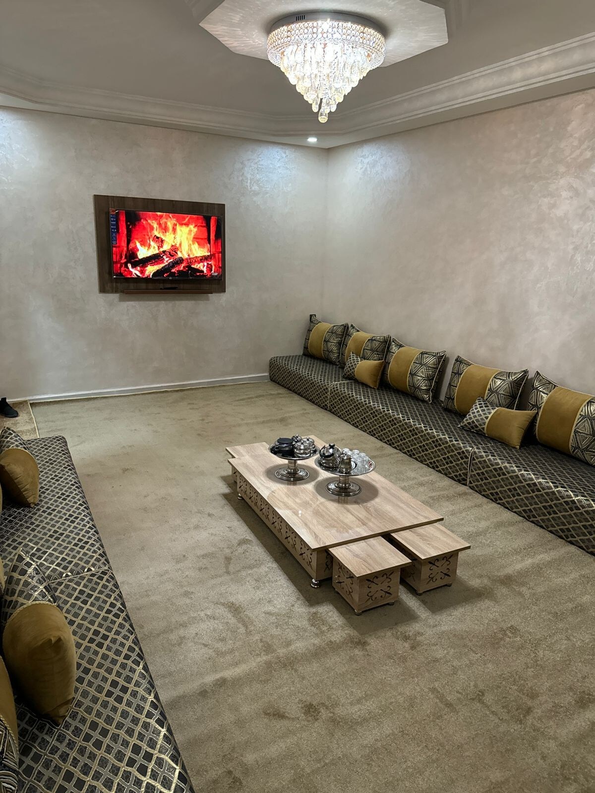 A spacious living room features a light-colored carpet and decorative low seating arranged around a central coffee table. An illuminated wall-mounted fireplace adds warmth, while elegant throw pillows enhance the seating area. Soft lighting is provided by a chandelier overhead.