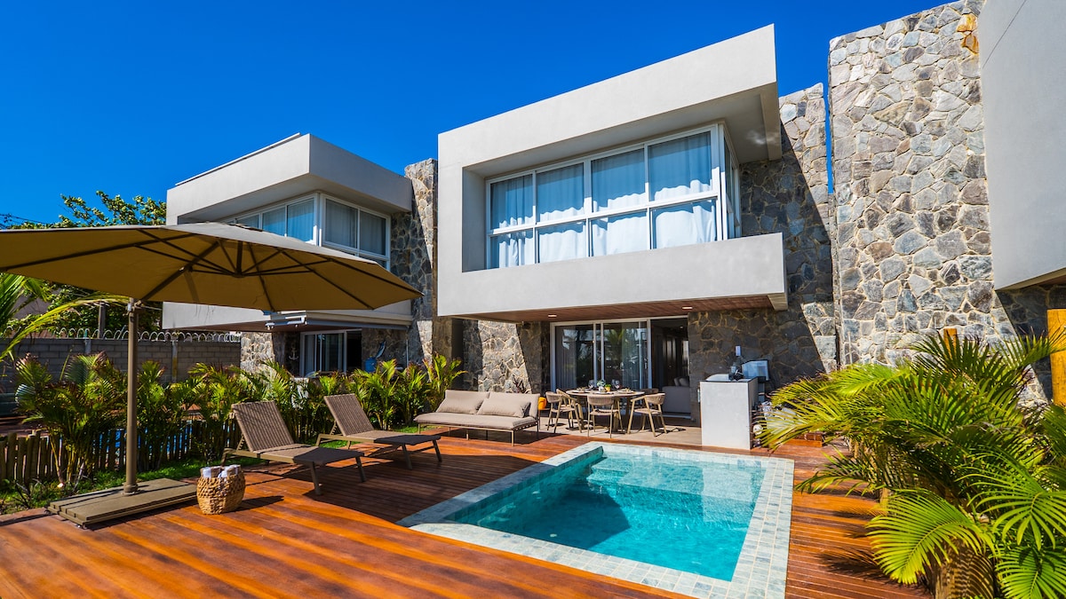 The exterior view displays a modern two-story house framed by natural stone and large windows. A private pool is situated in a wooden deck area, accompanied by loungers and a shaded umbrella. Lush tropical plants enhance the inviting outdoor space.