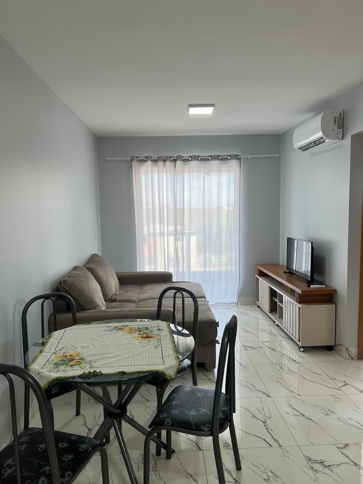 A light-filled living area is highlighted by a comfortable sofa and a small dining table set for four. A flat-screen television is positioned across from the seating, and sheer curtains frame a window that offers views of the outdoors.