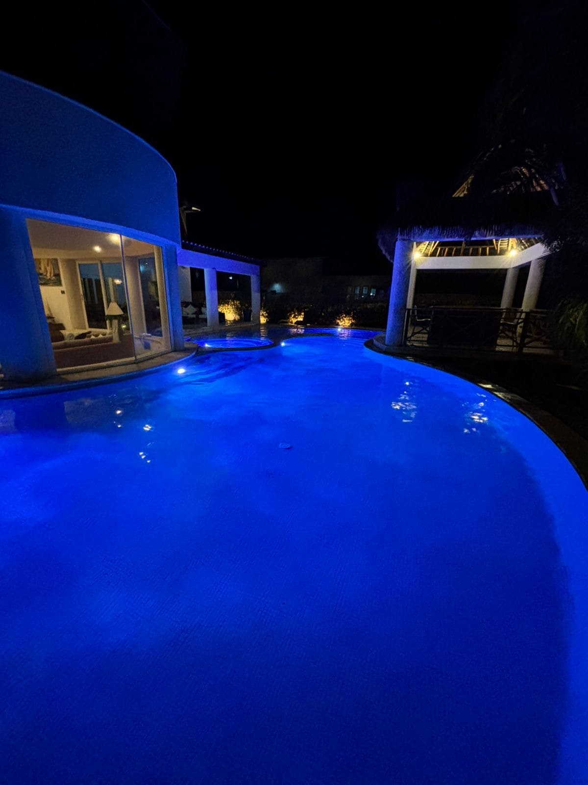 A curved pool is illuminated in soothing blue tones, reflecting the surrounding architecture. Soft lighting highlights the path alongside the pool, leading towards a cozy area with seating. In the background, the house is visible through expansive glass doors, showcasing an inviting interior.