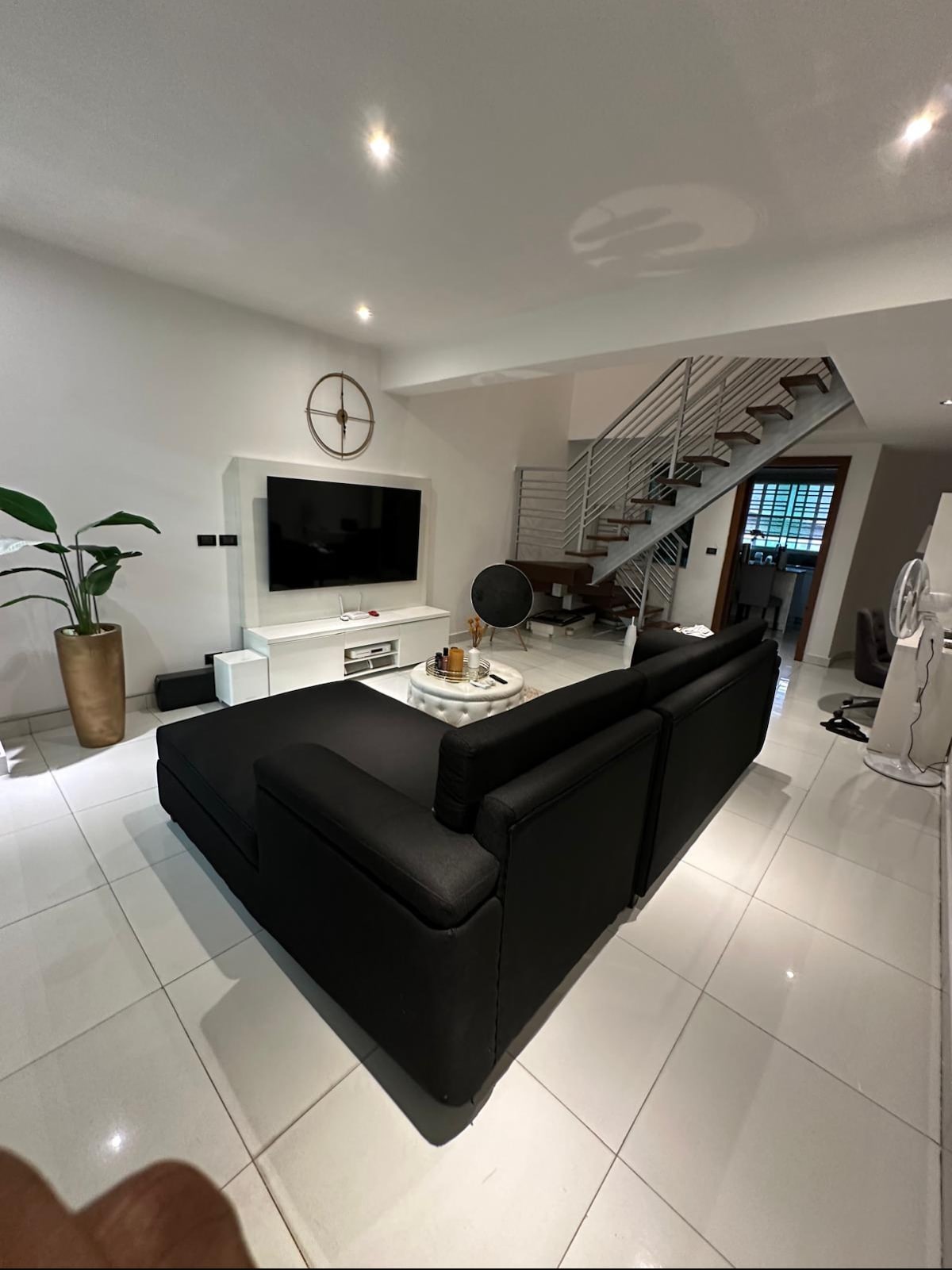 A modern living area features a sleek black sectional sofa positioned in front of a wall-mounted television. Light-colored tiles reflect soft overhead lighting, and a decorative plant adds a touch of greenery. An elegant staircase leads to an upper level.