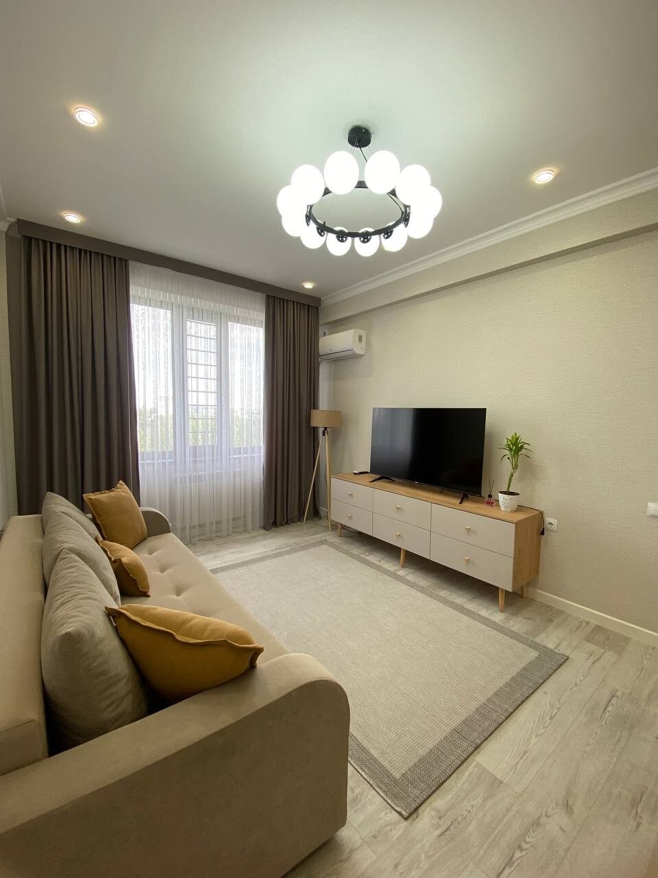 A light and airy living room features a comfortable beige sofa adorned with yellow cushions. A flat-screen television is placed on a light wooden console, complemented by a small green plant. Sheer curtains filter natural light from the adjacent window.