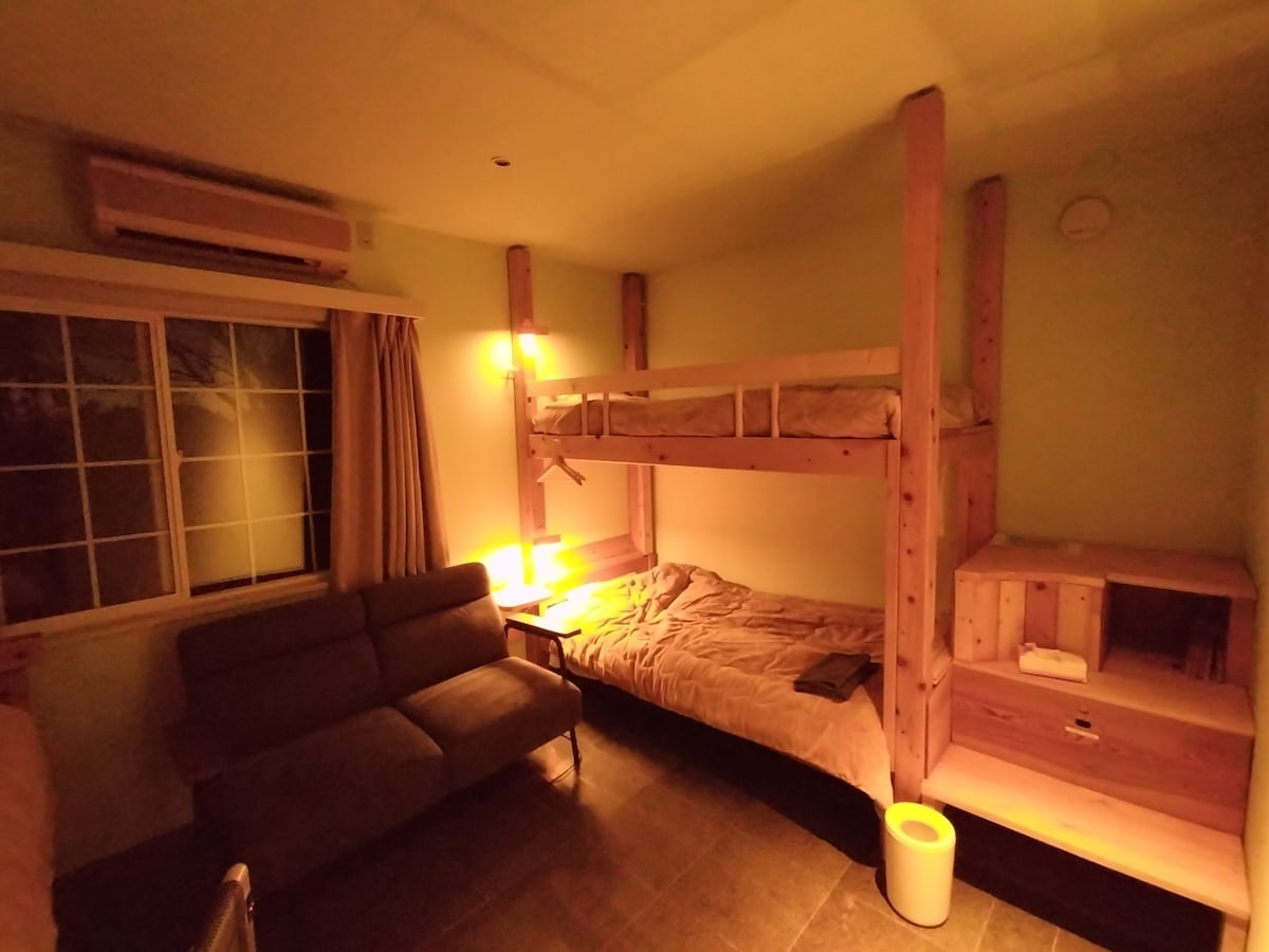 A mixed dormitory room features an upper bunk bed crafted from hinoki wood, illuminated by a reading light. A cozy lower bunk is present, along with a shared sofa area. Natural light filters through the large window, enhancing the serene ambiance.