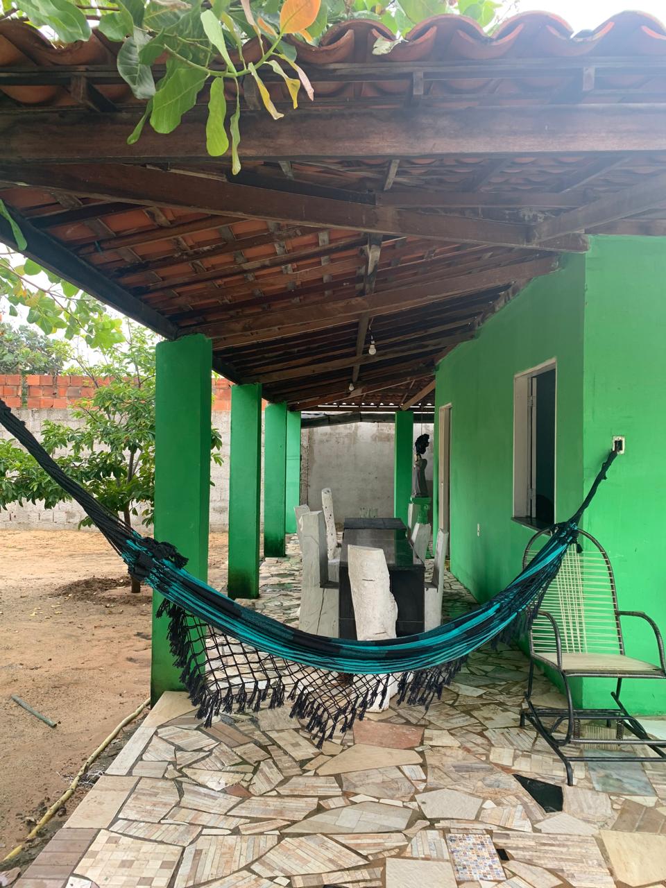 A covered patio area features a hanging hammock and a green accent wall. A series of stone tiles create the pathway, while a table and several chairs are positioned alongside. Natural light filters in through an uncovered roof, surrounded by lush greenery.