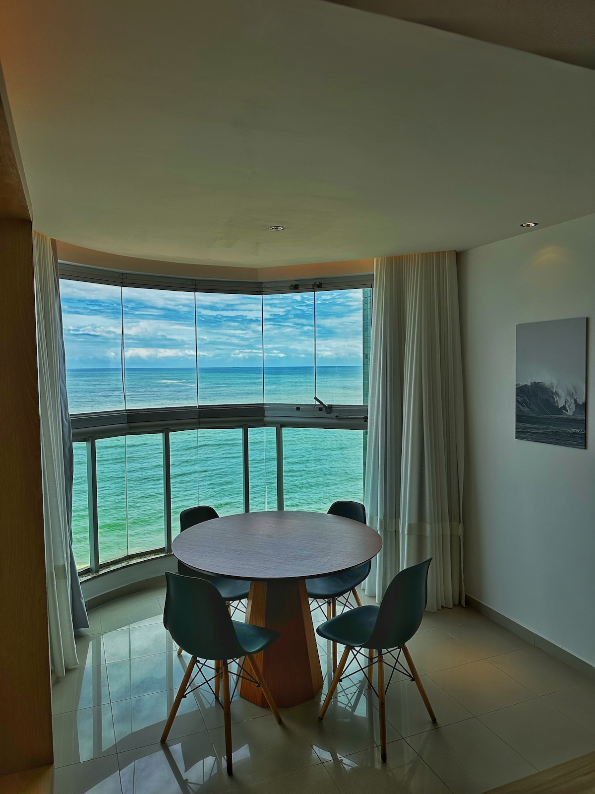 A round dining table surrounded by four chairs is positioned by a large curved window. Natural light floods the area, illuminating the space. The stunning view of the ocean and sky is prominently featured, framed by sheer white curtains.