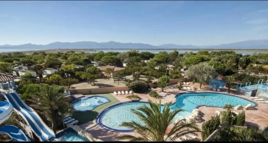 The image presents a spacious outdoor pool area surrounded by lush greenery and lounge chairs. Two pools, one with a waterslide, are visible. In the background, the tranquil lake and mountains create a serene setting, enhancing the overall view of the resort.
