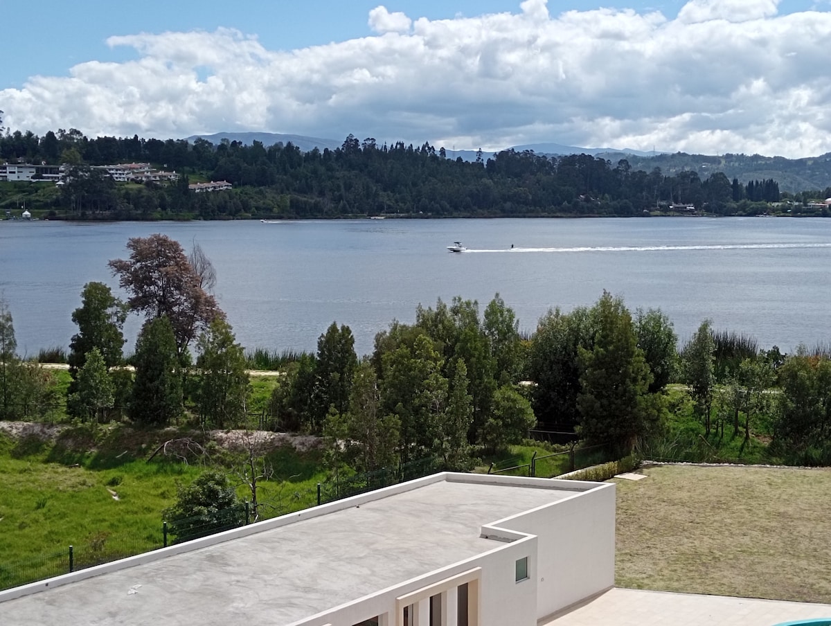 A serene view of the lake is captured, showcasing gentle waves and a boat gliding across the water. Lush greenery borders the lake, while rolling hills can be seen in the background, under a partly cloudy sky.