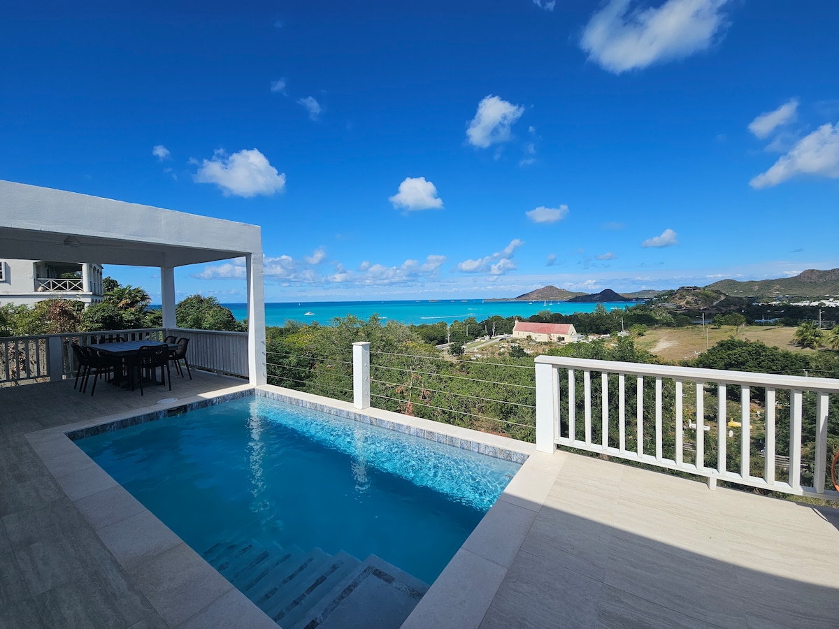 A private swimming pool sparkles under the sun, surrounded by a sleek deck with outdoor dining furniture. Scenic views of the Caribbean Sea and rolling hills create a vibrant backdrop, while the clear blue sky complements the tranquil surroundings.